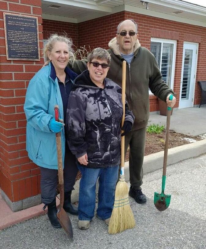 Three people stand outside a building holding gardening tools—a shovel, a broom, and another shovel. They appear to be engaged in outdoor work, smiling at the camera.