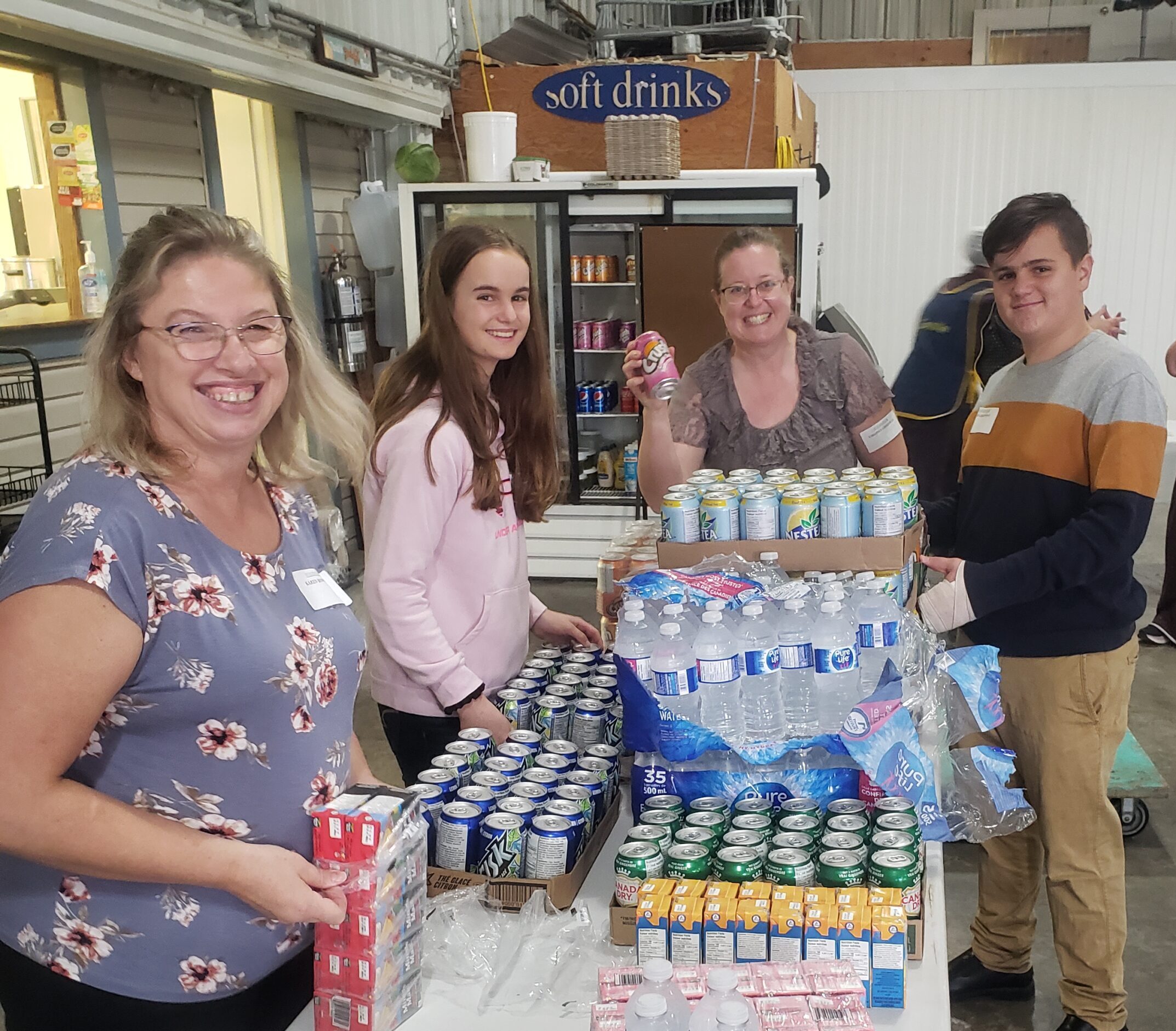 Four individuals are standing behind a table with stacks of canned soft drinks. They are smiling at the camera, possibly during a group activity or volunteering event.